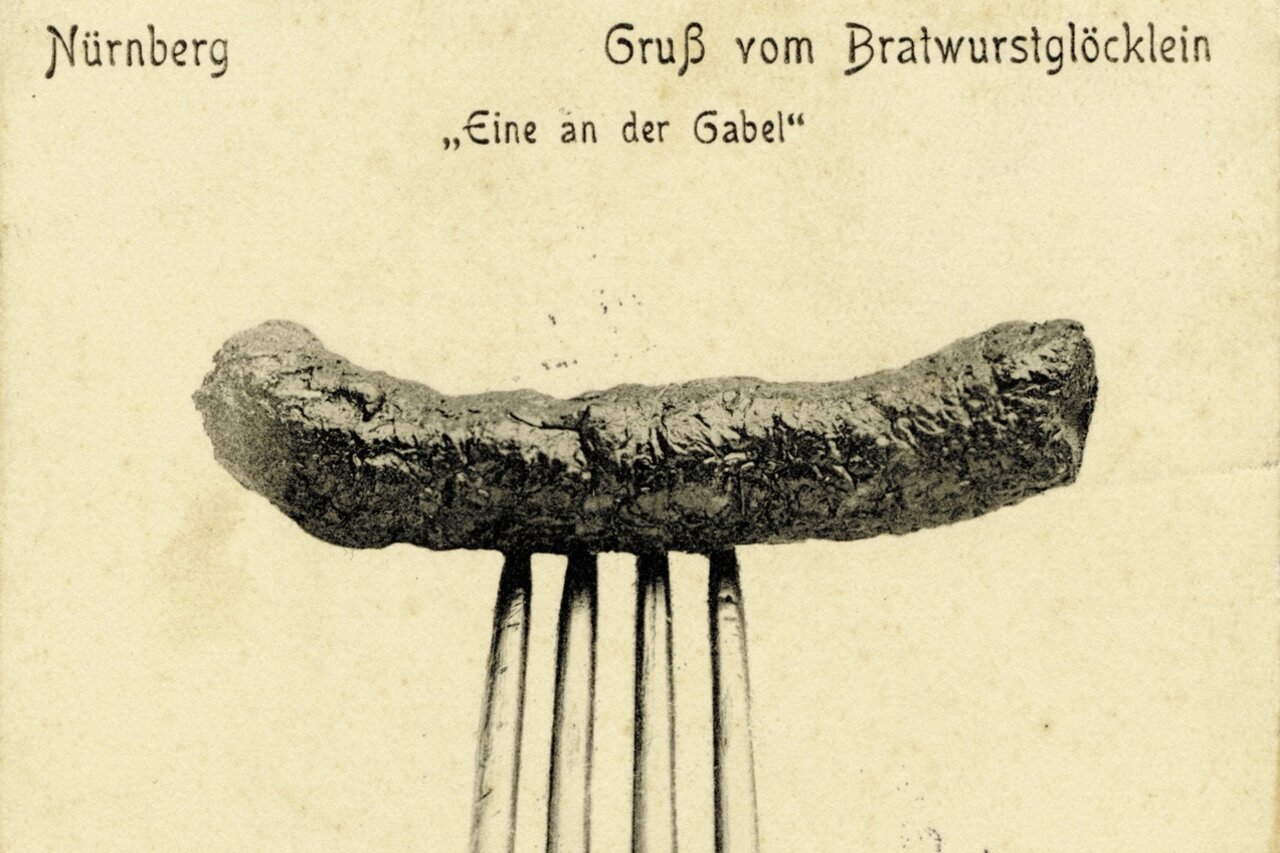 Historische Postkarte mit einer auf einer Gabel aufgespießten Bratwurst vor neutralem Hintergrund und der Aufschrift 'Nürnberg Gruß vom Bratwurstglöcklein Eine an der Gabel'