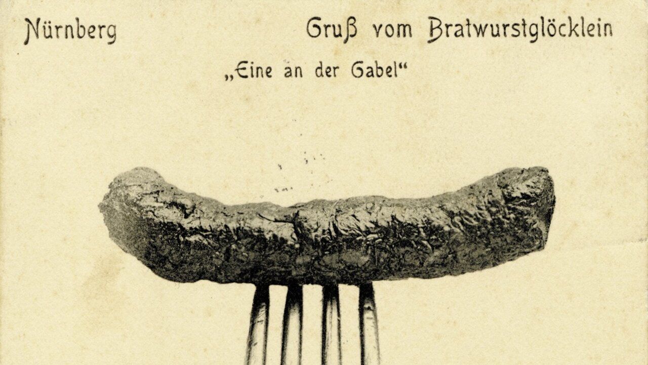 Historische Postkarte mit einer auf einer Gabel aufgespießten Bratwurst vor neutralem Hintergrund und der Aufschrift 'Nürnberg Gruß vom Bratwurstglöcklein Eine an der Gabel'