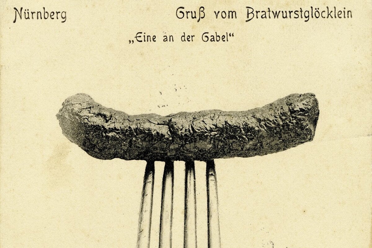 Historische Postkarte mit einer auf einer Gabel aufgespießten Bratwurst vor neutralem Hintergrund und der Aufschrift 'Nürnberg Gruß vom Bratwurstglöcklein Eine an der Gabel'
