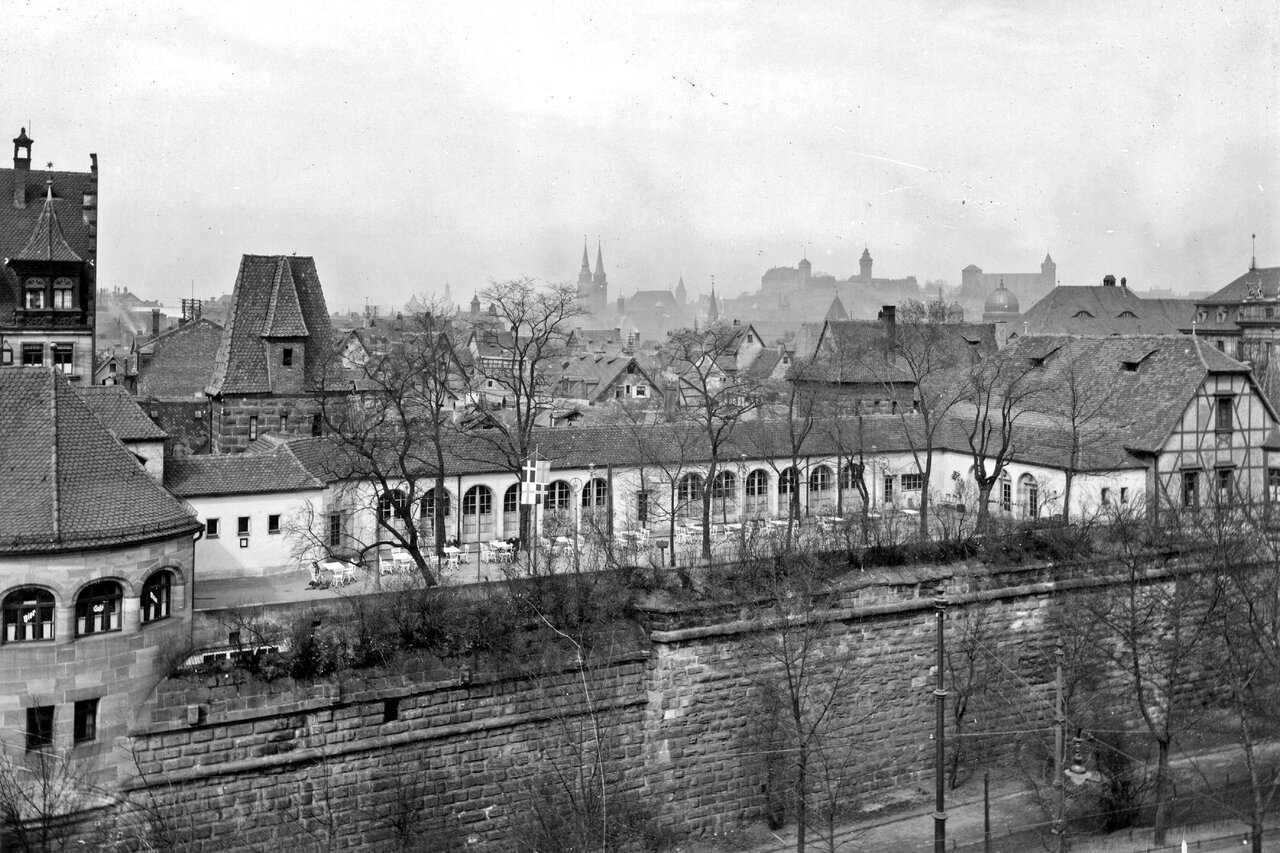 Schwarz-weiß Fotografie mit Gebäuden. Im unteren Bildteil die Stadtmauer, im Zwingerbereich eine Gaststätte. Hinter den Gastättengebäuden die Silhouette der Altstadt von Nürnberg, im Horizont die Kaiserburg.