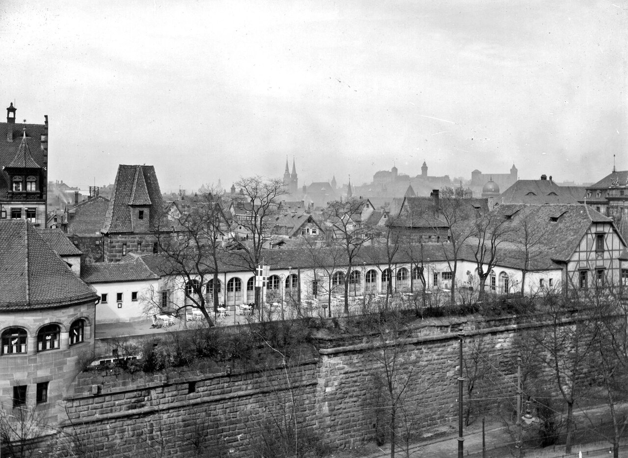 Schwarz-weiß Fotografie mit Gebäuden. Im unteren Bildteil die Stadtmauer, im Zwingerbereich eine Gaststätte. Hinter den Gaststättengebäuden die Silhouette der Altstadt von Nürnberg, im Horizont die Kaiserburg.