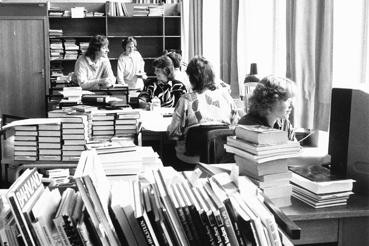 Schwarzweißaufnahme von einem Raum in einer Bibliothek, im Vordergrund Bücher, im Hintergrund sechs Personen, die sich mit den Büchern beschäftigen