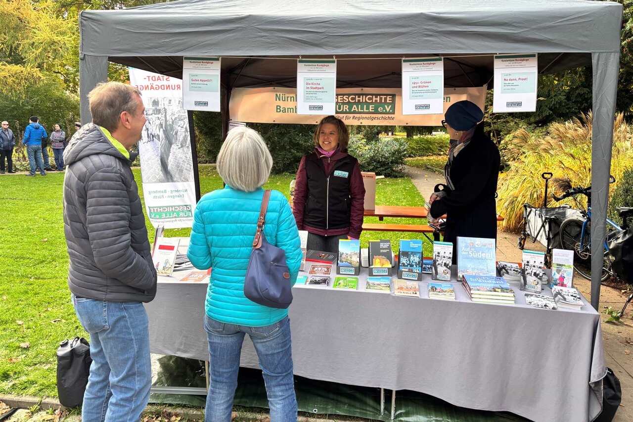Zwei Frauen stehen an einem Infostand, vor ihnen aufgereiht auf einem Tisch Bücher und Flyer von Geschichte Für Alle, ein Frau und ein Mann sind mit ihnen im Gespräch.