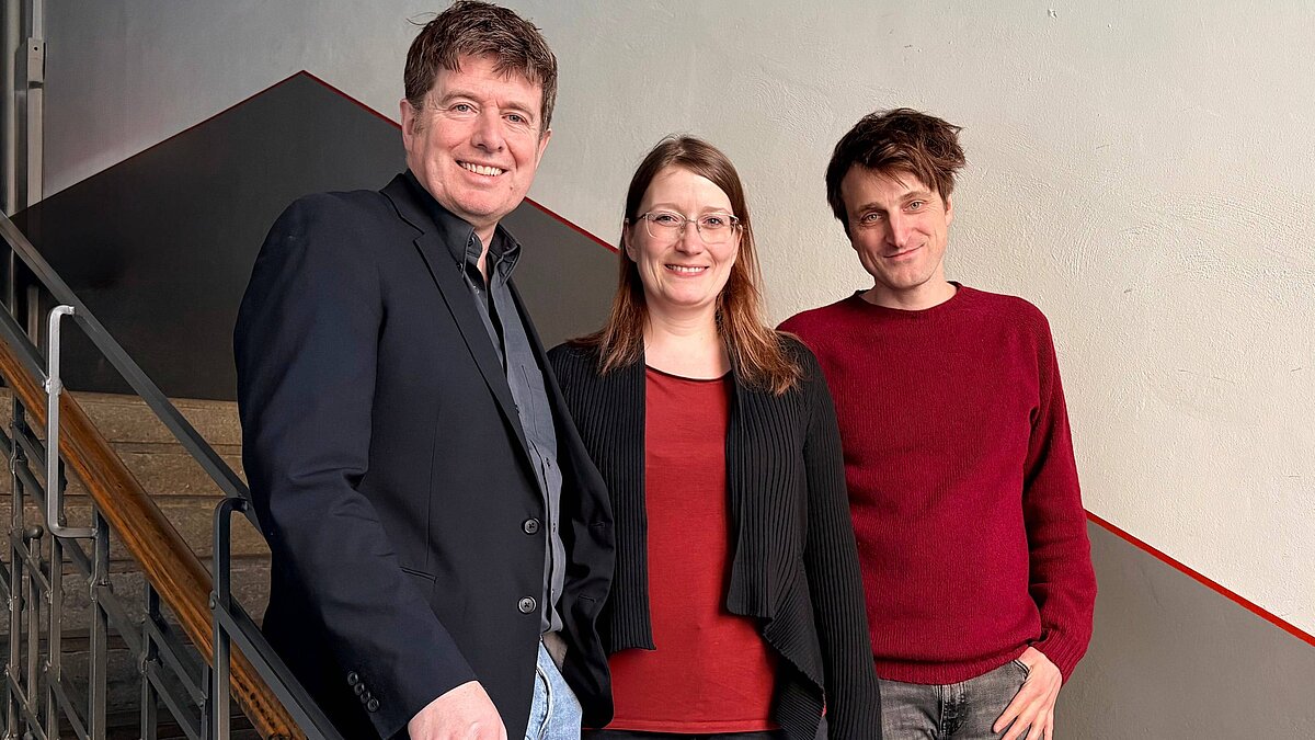 Auf dem Bild sind drei Personen zu sehen, von links nach rechts: ein Mann, lächelnd, mit dunklem Sakko; lächelnde Frau in der Mitte, roter Pullover, schwarze Jacke; lächelnder Mann mit rotem Pullover