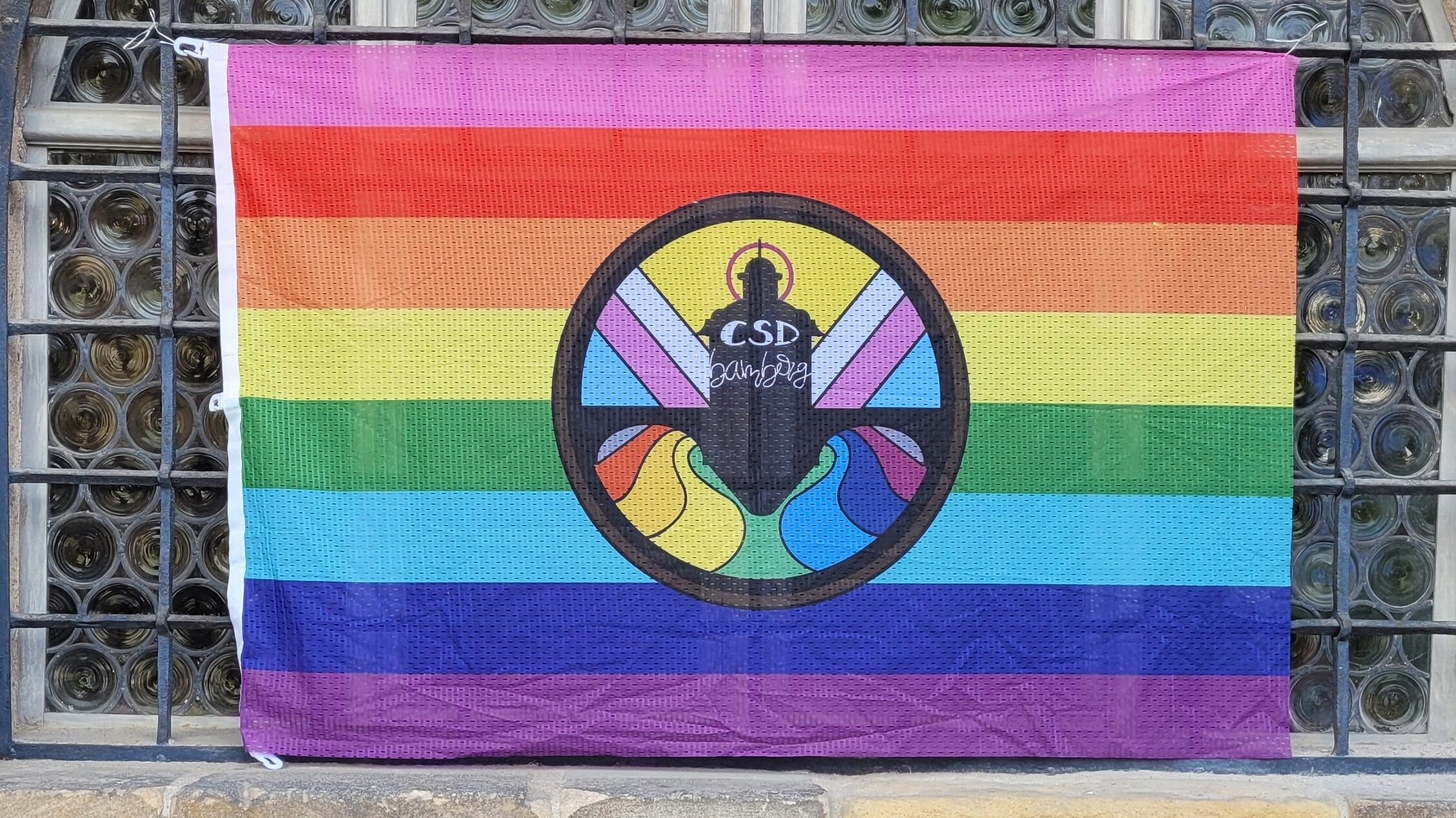 Fotografie einer Regenbogenfahne, die an einem Gitter vor einem Fenster hängt. In der Mitte der Fahne ein buntes Logo mit einer Gebäudeilhouette, auf dieser steht die Schrift "CSD Bamberg". 