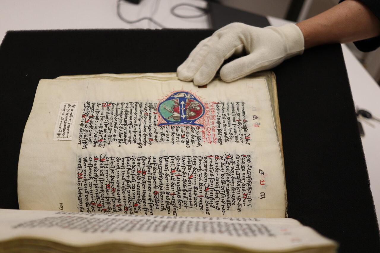 Ein altes Buch mit Frakturschrift und bunten Malereien liegt offen auf einem schwarzen Untergrund und wird von einer Hand mit weißem Handschuh offen gehalten