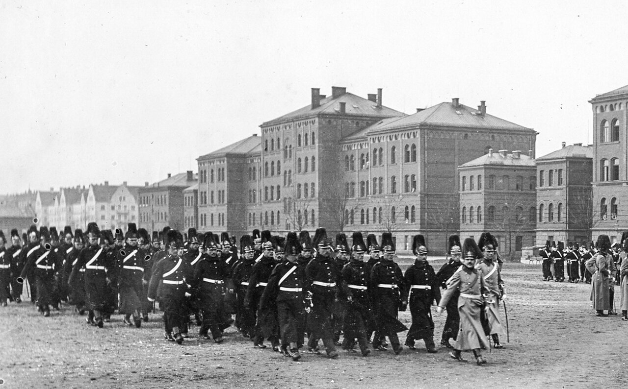Schwarzweißaufnahme von marschierenden Soldaten in schwarzen Uniformen auf einer großen Straße, im Hintergrund große Steingebäude