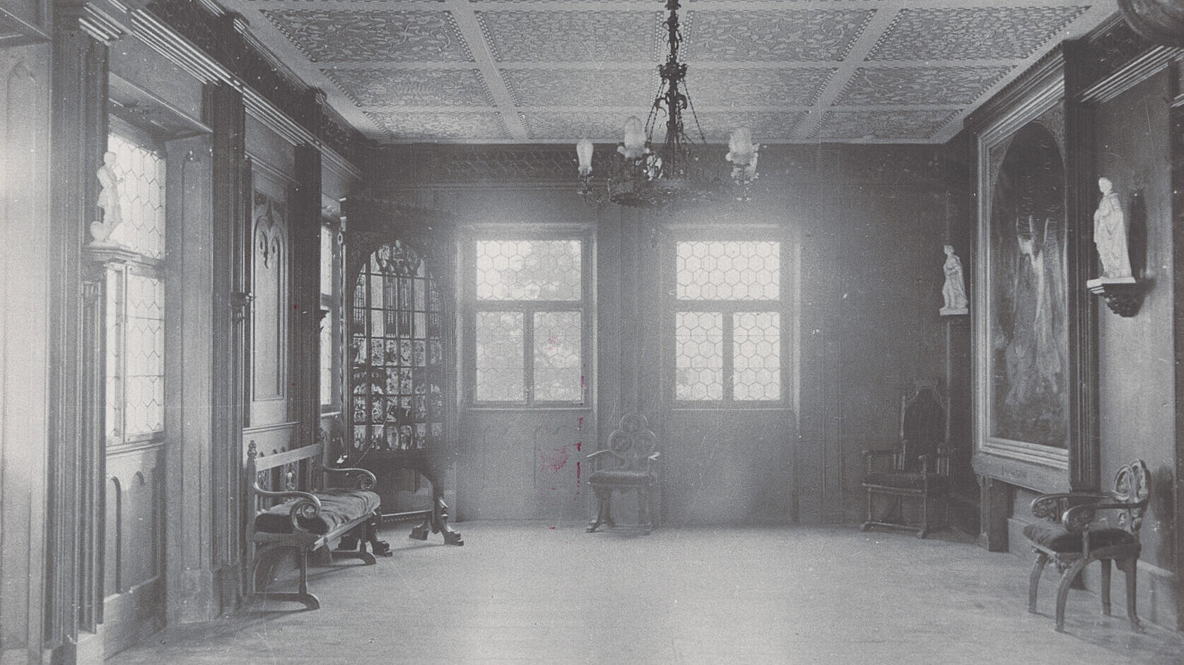 Schwarzweißaufnahme eines Saals mit großen Fenstern, Kronleuchtern, Gemälden und verzierten Holzmöbeln, um 1900