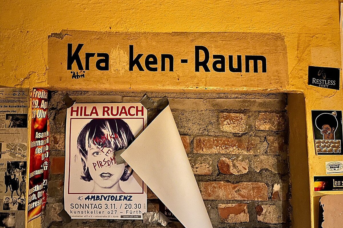 Plakate im kunstkeller o27 © Claudia Oehm, 2025. Wand im Luftschutzkeller mit bunten Veranstaltungsplakaten und Stickern und einem alten zugemauerten Türstock über dem "Krnkenraum" geschrieben steht.