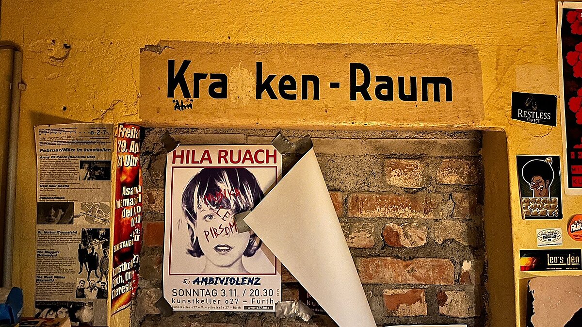 Plakate im kunstkeller o27 © Claudia Oehm, 2025. Wand im Luftschutzkeller mit bunten Veranstaltungsplakaten und Stickern und einem alten zugemauerten Türstock über dem "Krnkenraum" geschrieben steht.