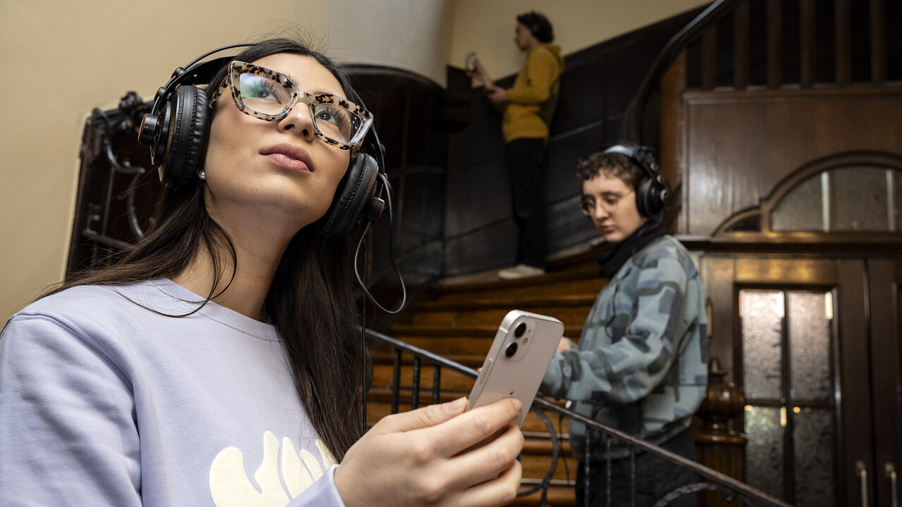 Junge Frau mit Kopfhörern und Smartphone in der Hand schaut sich interessiert um, im Hintergrund zwei weitere junge Menschen mit Kopfhörern im Treppenhaus eines Alten Gebäudes