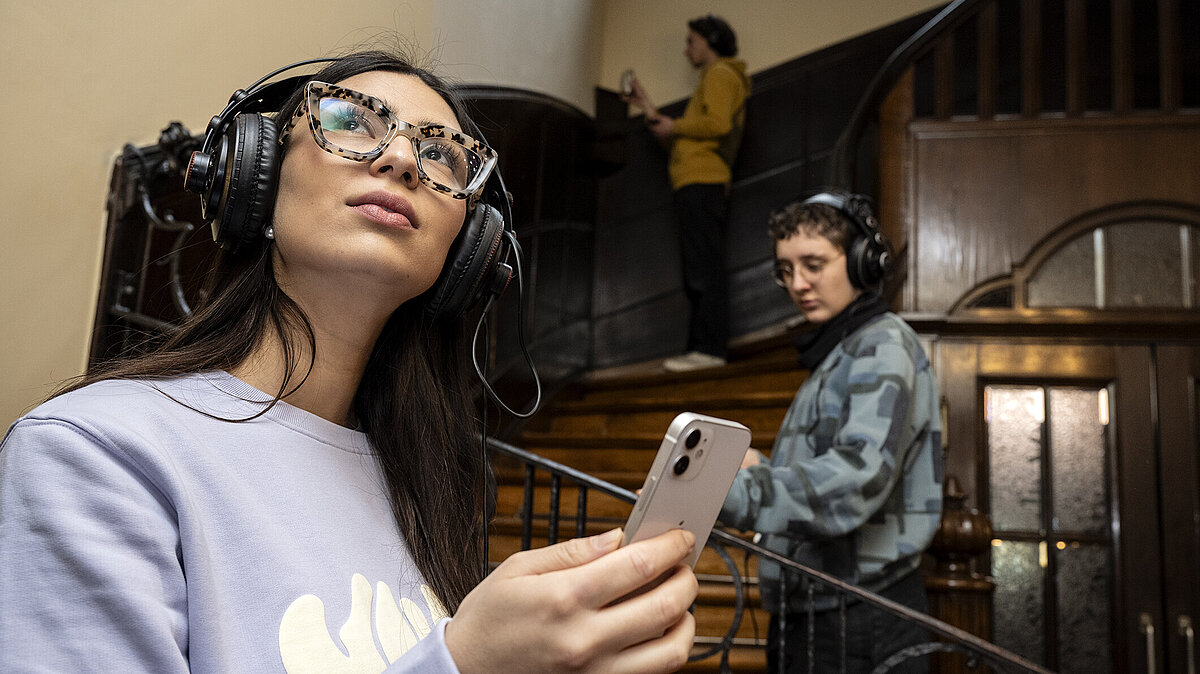 Junge Frau mit Kopfhörern und Smartphone in der Hand schaut sich interessiert um, im Hintergrund zwei weitere junge Menschen mit Kopfhörern im Treppenhaus eines Alten Gebäudes