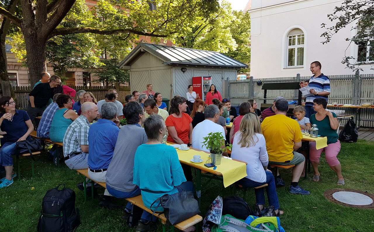 Eine Gruppe Menschen sitzt an Gartentischen und hört einem Redner zu, im Hintergrund eine graue Gartenhütte und Bäume