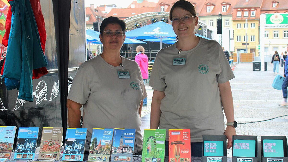 Franca Heinsch und Ulla Hoßfeld am Infostand in Bamberg © Amely Wild, 2025. Zwei Frauen stehen an einem Infostand, vor ihnen aufgereiht auf einem Tisch Bücher und Flyer von Geschichte Für Alle.