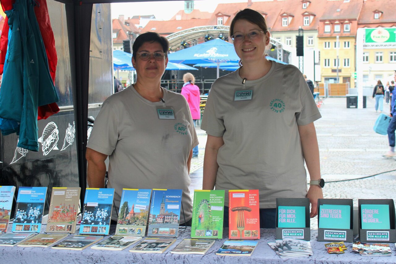Zwei Frauen stehen an einem Infostand, vor ihnen aufgereiht auf einem Tisch Bücher und Flyer von Geschichte Für Alle.
